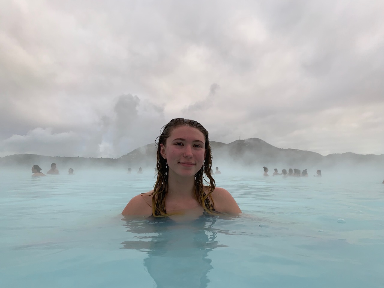 Milky-blue geothermal lagoon with bathers among dark volcanic rocks under a bright sky.