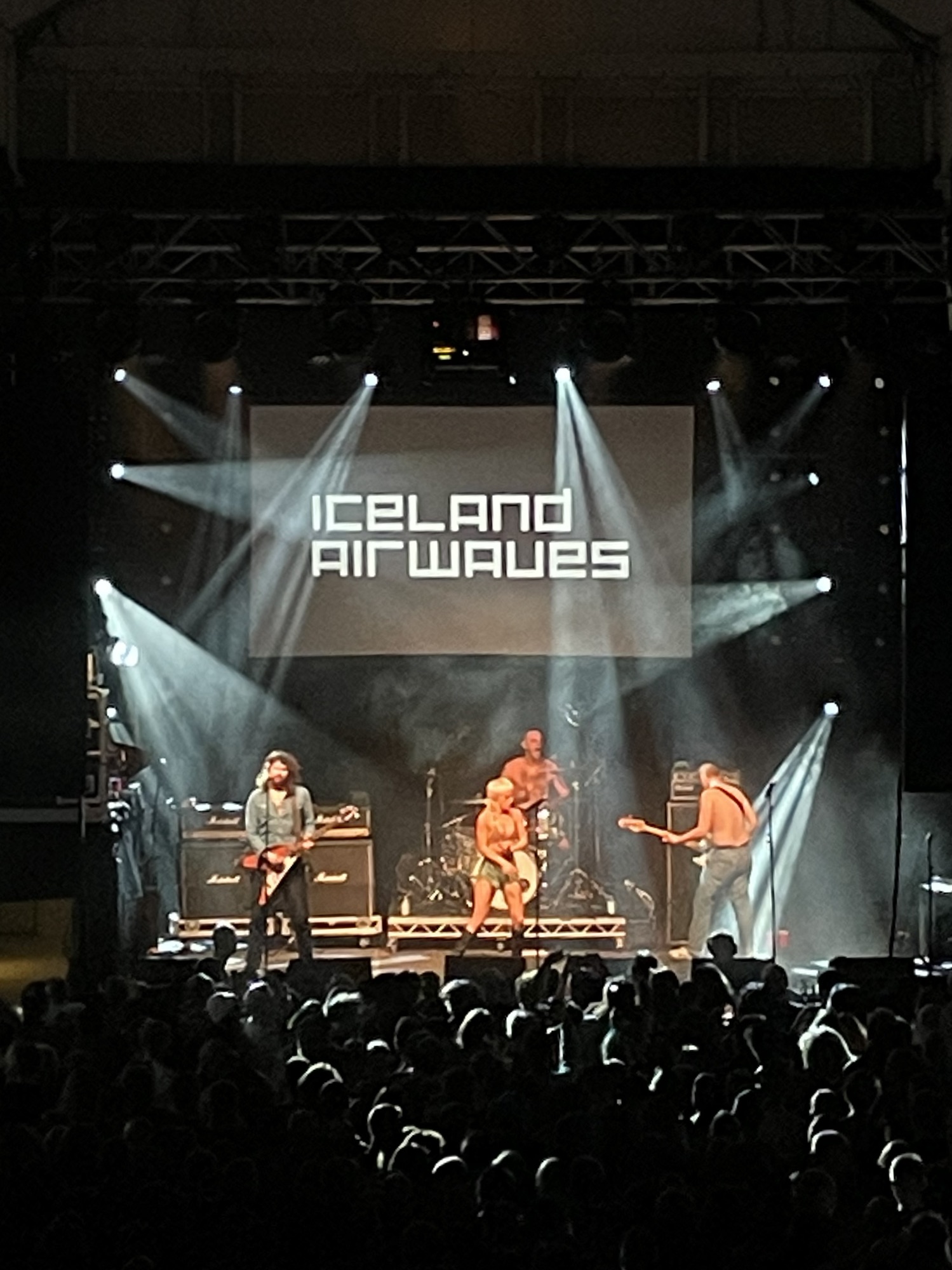 A four-piece band performing on stage at Iceland Airwaves with the festival name projected on the screen behind them and a crowd in front.