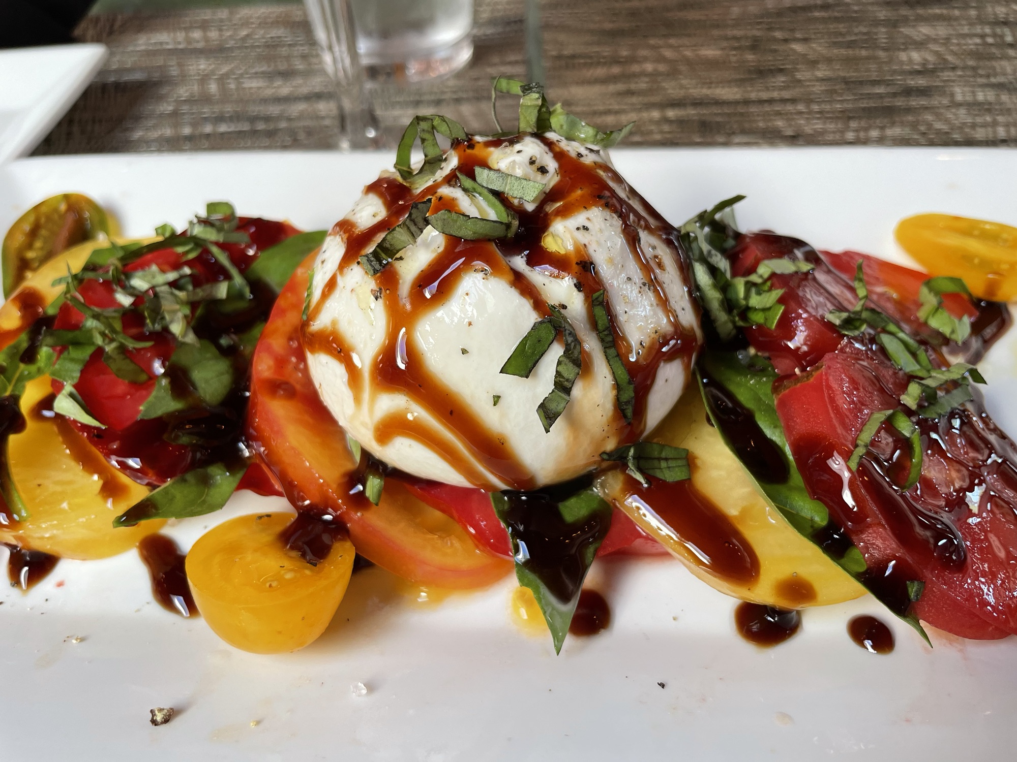 A ball of burrata centered on heirloom tomato slices with basil and a balsamic drizzle on a white plate.